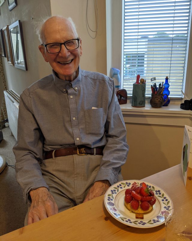 A smiling man with grey hair and glasses sitting in front of a plate of fruit and pancakes