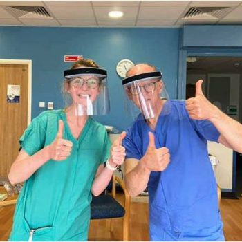 a man and a woman wearing scrubs giving the thumbs up sign while wearing face visors
