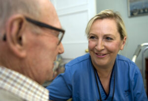 Image shows a nurse in a blue uniform with an elderly man in glasses. Both are smiling