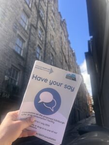 Image shows a hand holding a leaflet on the safter drug consumption facility. It is taken outside with Edinburgh Old Town in the background