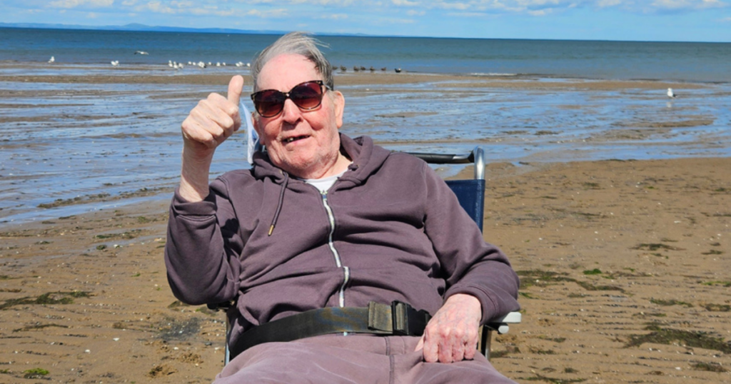 A resident in a wheelchair on the beach gives a thumbs up